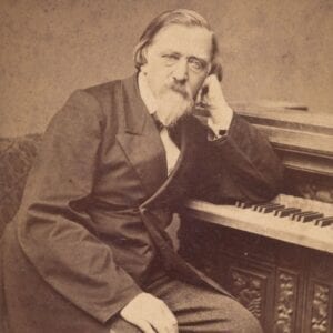 Sepijos atspalvio XIX a. portretinė fotografija: vyresnio amžiaus vyras su barzda sėdi prie pianino