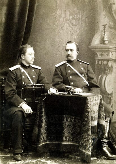 Rusijos imperijos karininkai XIX a. nuotraukoje Du kariniais mundurais vilkintys vyrai fotoateljė prie kilimu užtiesto stalelio, senovinė nespalvota nuotrauka